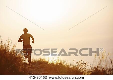 Stock Image - Man running on trail overlooking sunset ocean. Fotosearch