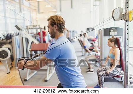Stock Image - Man using cable exercise equipment at gym. Fotosearch