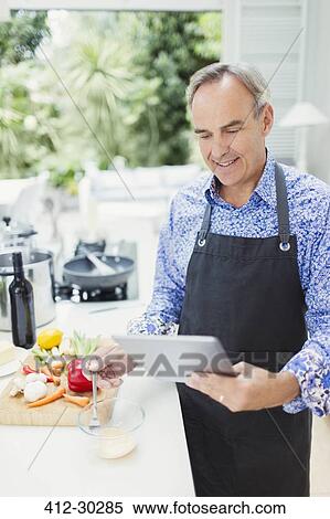 Stock Photography - Mature man in apron using digital tablet cooking in kitchen. Fotosearch
