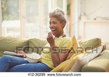 Stock Photo - Mature woman eating popcorn and watching TV on living room sofa. Fotosearch