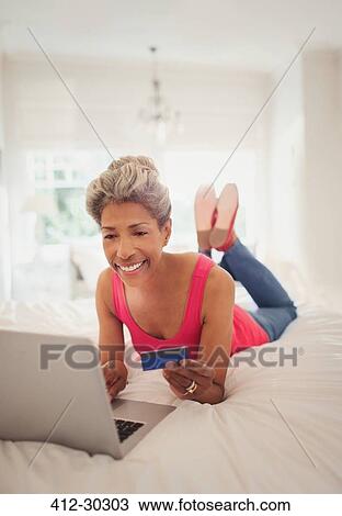 Mature woman with credit card shopping online with laptop on bed View Large Photo Image Stock Image - Mature woman with credit card shopping online with laptop on bed. Fotosearch