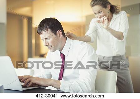 Stock Image - Mischievous businesswoman aiming rubber band at unsuspecting businessman working at laptop. Fotosearch
