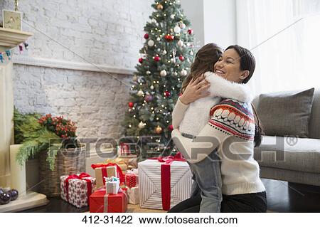 Stock Image - Mother and daughter hugging near Christmas tree and gifts in living room. Fotosearch