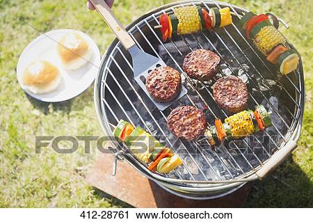Stock Image - Overhead view of hamburgers and vegetable skewers on barbecue grill. Fotosearch