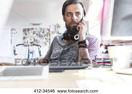Stock Photograph - Pensive design professional with headphones talking on cell phone at desk. Fotosearch