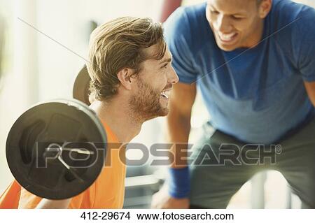 Picture - Personal trainer encouraging man doing barbell squats at gym. Fotosearch