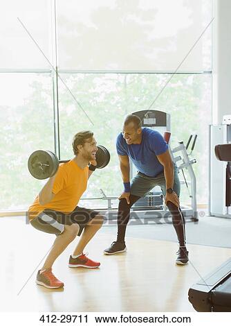 Stock Image - Personal trainer guiding man doing barbell squats at gym. Fotosearch