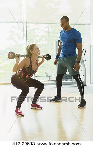 Stock Image - Personal trainer guiding woman doing barbell squats at gym. Fotosearch