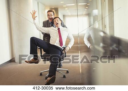 Stock Photography - Playful businessman pushing colleague down corridor in office chair. Fotosearch