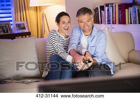 Playful couple fighting over remote control watching TV in living room View Large Photo Image Picture - Playful couple fighting over remote control watching TV in living room. Fotosearch