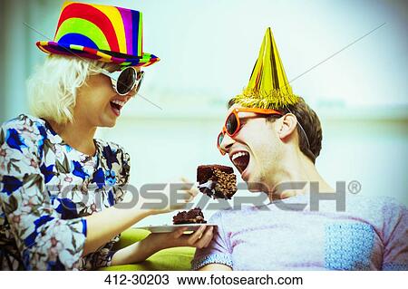 Stock Image - Playful couple in party hats and sunglasses eating chocolate cake. Fotosearch