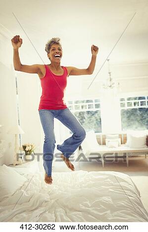 Stock Image - Playful enthusiastic mature woman jumping on bed. Fotosearch