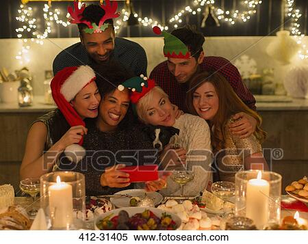 Stock Photography - Playful friends with dog taking selfie at candlelight Christmas table. Fotosearch