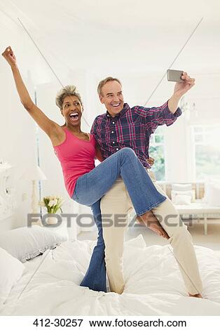 Stock Photo - Playful mature couple taking selfie standing on bed. Fotosearch