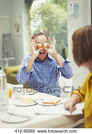 Stock Photography - Playful mature man covering eyes with eggs at breakfast table. Fotosearch