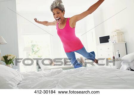 Picture - Playful mature woman jumping onto bed. Fotosearch