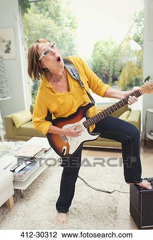 Stock Image - Playful mature woman playing electric guitar in living room. Fotosearch