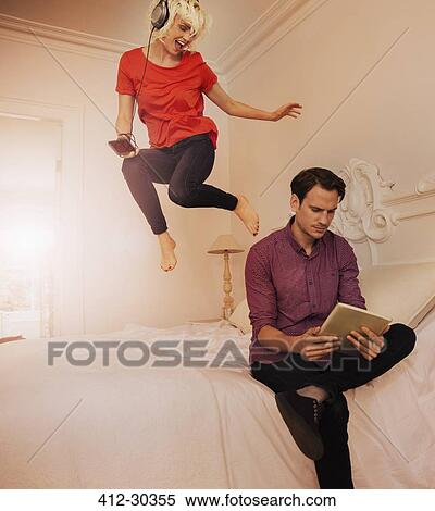 Stock Photography - Playful wife jumping on bed listening to music behind husband using digital tablet. Fotosearch
