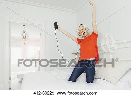 Stock Photography - Playful women kneeling on bed listening to music with mp3 player and headphones. Fotosearch