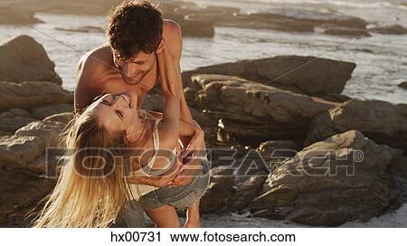 Stock Image - Playful young couple hugging on rocks at ocean. Fotosearch