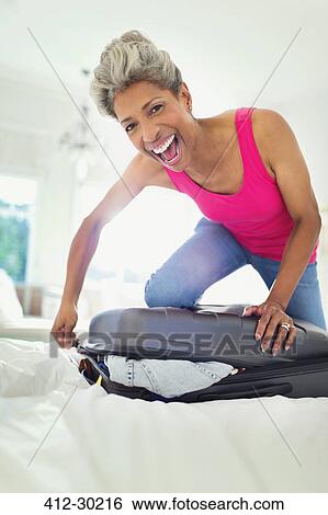 Stock Photograph - Portrait enthusiastic mature woman closing suitcase on bed. Fotosearch