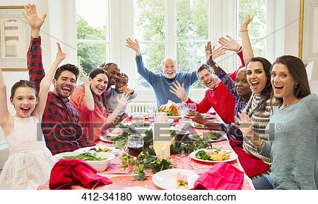 Stock Image - Portrait enthusiastic multi-ethnic multi-generation family waving at Christmas dinner table. Fotosearch