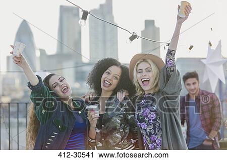 Stock Image - Portrait exuberant young women cheering and drinking at rooftop party. Fotosearch