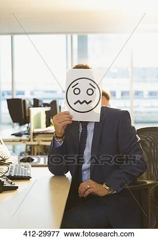 Stock Photo - Portrait of businessman holding frowning face printout over his face in office. Fotosearch