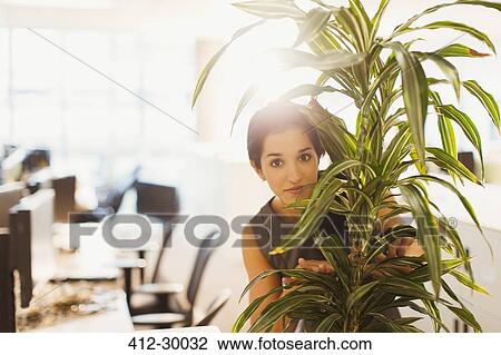 Stock Image - Portrait of businesswoman hiding behind plant in office. Fotosearch