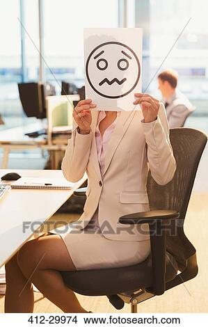 Portrait of businesswoman holding frowning face printout over her face in office View Large Photo Image Picture - Portrait of businesswoman holding frowning face printout over her face in office. Fotosearch