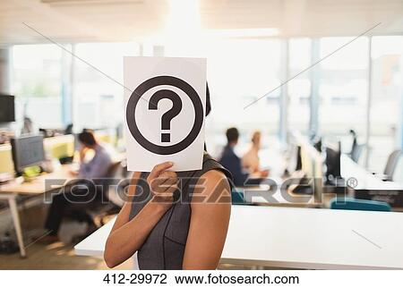 Stock Image - Portrait of businesswoman holding question mark printout over her face in office. Fotosearch