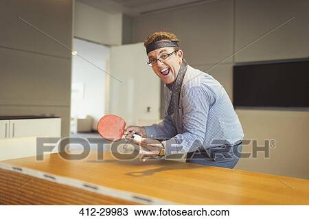 Stock Image - Portrait of enthusiastic businessman playing ping pong with tie wrapped around head in conference room. Fotosearch