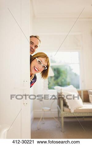 Stock Image - Portrait playful mature couple peering from behind door. Fotosearch