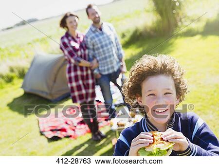 Portrait smiling boy eating hamburger with parents at sunny campsite View Large Photo Image Stock Image - Portrait smiling boy eating hamburger with parents at sunny campsite. Fotosearch