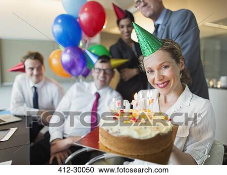 Stock Image - Portrait smiling businesswoman holding birthday cake with colleagues in background. Fotosearch