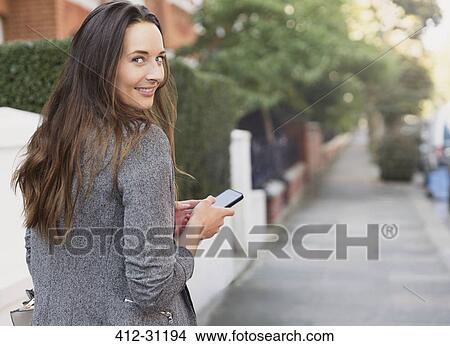 Picture - Portrait smiling businesswoman with cell phone looking back on sidewalk. Fotosearch