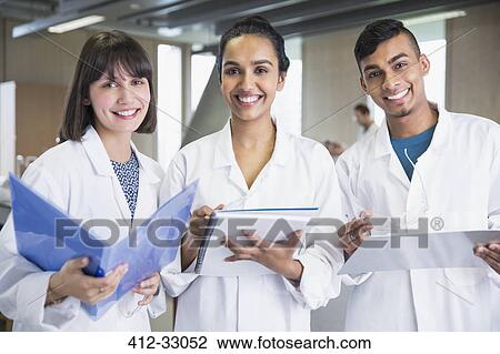 Stock Image - Portrait smiling college students in lab coats with notebooks in science laboratory classroom. Fotosearch