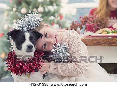Stock Photograph - Portrait smiling girl hugging dog with Christmas decoration. Fotosearch