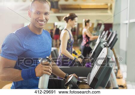 Stock Image - Portrait smiling man with water bottle on treadmill at gym. Fotosearch
