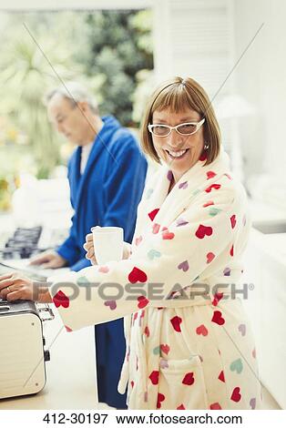 Stock Photo - Portrait smiling mature woman drinking coffee in bathrobe at kitchen counter. Fotosearch