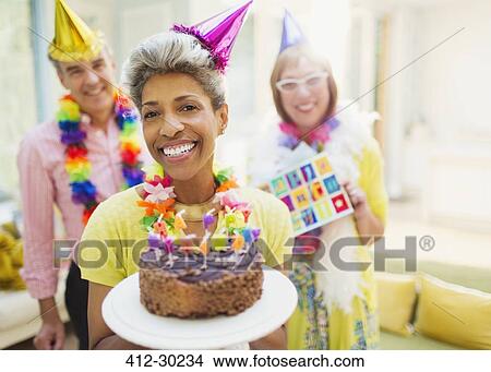 Picture - Portrait smiling mature woman holding chocolate birthday cake with friends in background. Fotosearch
