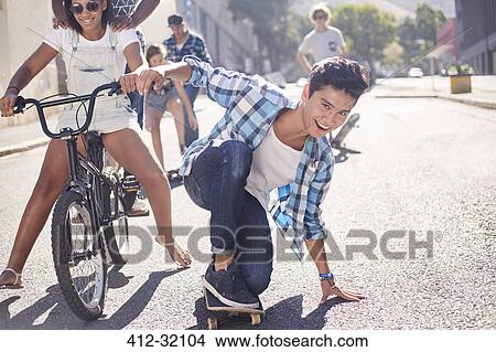 Picture - Portrait smiling teenage boy skateboarding with friends on sunny urban street. Fotosearch