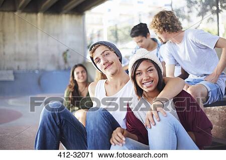 Stock Image - Portrait smiling teenage couple hanging out with friends at skate park. Fotosearch