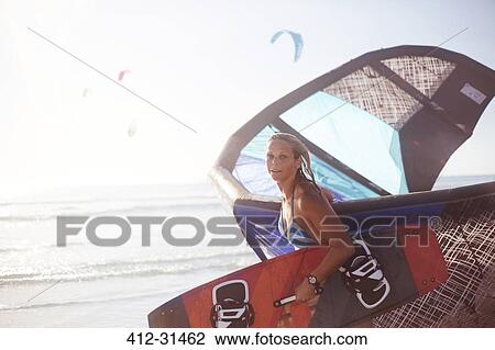 Stock Image - Portrait woman carrying kiteboard equipment on beach. Fotosearch