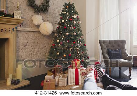 Stock Photograph - Relaxed couple wearing socks with feet up near Christmas tree in living room. Fotosearch