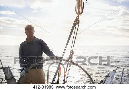 Stock Image - Retired man sailing steering sailboat at helm. Fotosearch