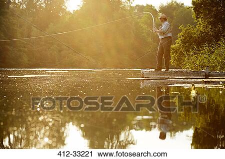 Stock Image - Senior man fly fishing on dock of summer lake. Fotosearch