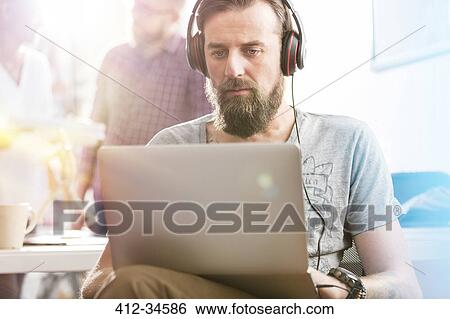 Stock Photograph - Serious male design professional with headphones using laptop in office. Fotosearch
