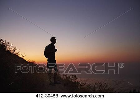 Stock Photo - Silhouette of male hiker on trail overlooking ocean at sunset. Fotosearch