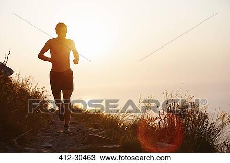 Stock Image - Silhouette of man running on trail with sunset ocean in background. Fotosearch